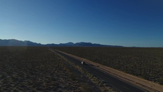 An aerial, tracking shot of a moving vehicle coming to the road split in a vast valley, with grandiose mountains visible in the background