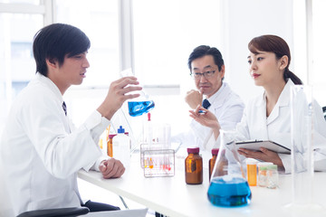 フラスコの中の青い液体を見つめながら議論をする三人の研究員たち