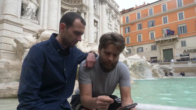 Male Tourist Looking At Smartphone Screen Sat In Front Of Trevi Fountain.