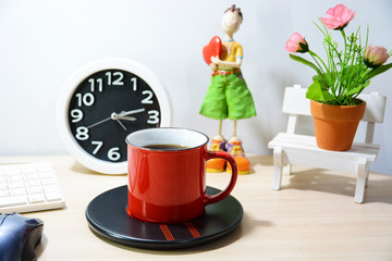 Red coffee mug Put on the desk Have a doll watch and a flower vase next to it.
