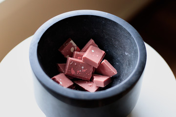 Broken and shattered pieces of ruby red chocolate in a round charcoal stone ceramic bowl. White and brwon background. Speckled chocolate.