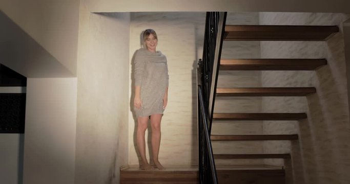 Wide shot of a beautiful blonde woman with bare legs beckoning the viewer to come up stairs