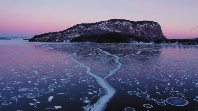 Flying low and slow at dawn over the cracks in a frozen lake towards the cliff face of an isolated mountain
