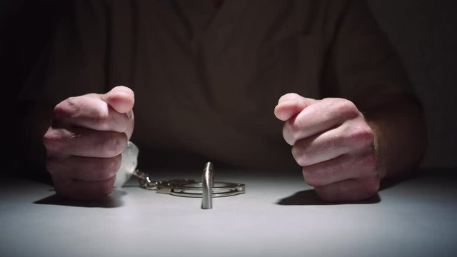 Close-up of a prisoner's hands while he sits calmly handcuffed to a table in an interrogation room, his hands balled into fists.