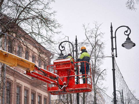 Workers in lift bucket during installation festive luminous garlands. Winter evening illumination. Worker in lift bucket repair street light pole with double head lamps. Modernization of street lamps