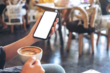 Mockup image of hands holding white mobile phone with blank screen while drinking coffee in modern cafe