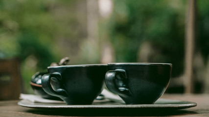 Two black cups of coffee on a table a coffee shop, with green plants on the background.