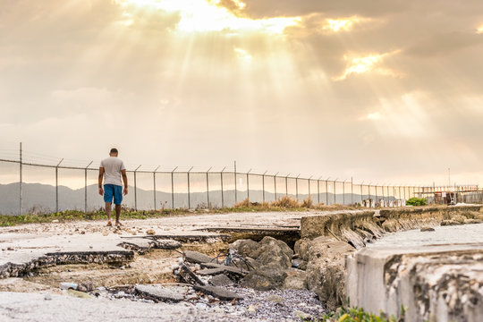 Broken Young Black Man Walks On Road By Sea/ Ocean Water Coast Toward Sun Rays Beaming Through Clouds. Journey Into The Unknown. Leave Troubles Behind. Walking Away From Past. Life Decisions/ Choices.