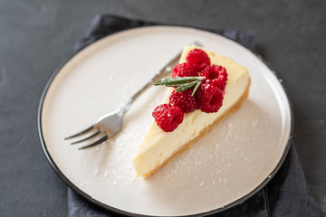 Slice of New York Cheesecake with raspberries on a dark background.