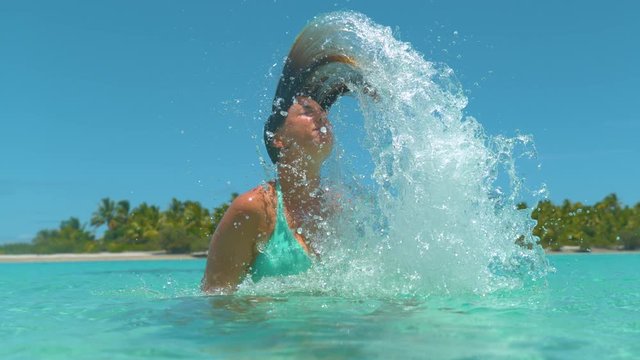 SLOW MOTION, CLOSE UP: Playful Caucasian woman on carefree holiday whips her wet hair back and sprays the glassy ocean water. Happy tourist girl splashing the sea water with her long brown hair.