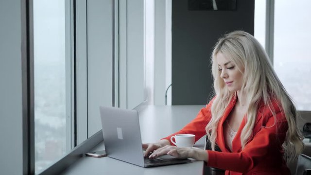 Woman in red coral business suit. Young blonde businesswoman with modern gray laptop in the office in high floor in front of the window. Girl drinking coffee
