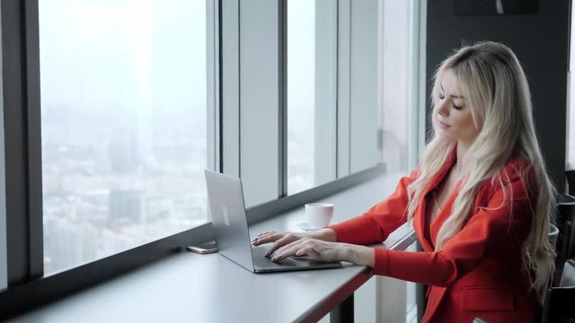 Woman in red coral business suit. Young blonde businesswoman with modern gray laptop in the office in high floor in front of the window. Girl drinking coffee