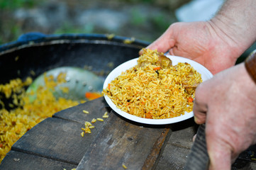 Human hands and a portion of pilaf in a white plastic plate on the Board lid of the cauldron. Background. Lunch for a large group of tourists