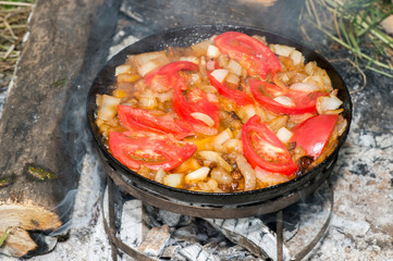 Tomatoes are fried on pork fat on a black pan on a metal grill during the journey for the preparation of scrambled eggs