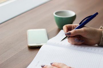 Close up of woman's hand writing something in a notebook on the background of a Cup, a phone and a wooden table. An unrecognized business woman is planning her day. Planning, work, business concept.
