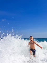 Fototapeta premium A little blond boy is playing on the seashore, laughing, running away from the waves against the background of huge waves and splashes, Calabria, Italy