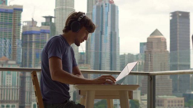 Young man freelancer workes on his laptop at a balcony with a background of a city center full of skyscrapers. Remote techer has a conversation with a student through the internet