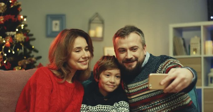Close up of the cheerful parents and cute son sitting on the couch at the x-mas tree in the living room and taking selfie picture on the smartphone camera.