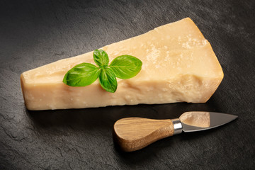 A piece of aged Parmesan cheese on a black background with fresh basil leaves, a cheese knife, and a place for text