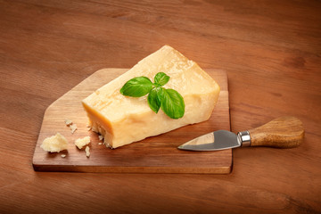 A piece of aged Parmesan cheese with crumbs, fresh basil and a cheese knife on a dark rustic wooden background with a place for text
