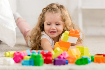 happy adorable kid playing with colored plastic constructor on carpet in children room