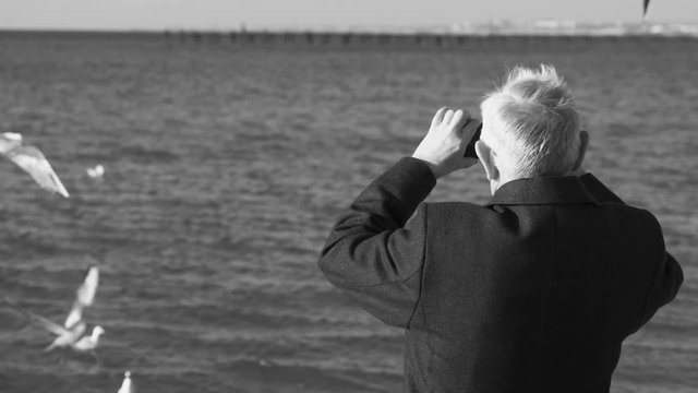 Elderly gray-haired man photographed on a mobile phone seascape with a pier and seagulls. Back view.