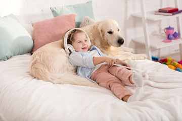adorable kid listening music with tablet and leaning on fluffy golden retriever on bed in children room