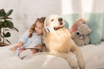 adorable child holding tablet, cute golden retriever with headphones lying on bed in children room