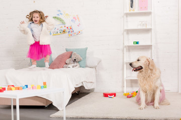 excited adorable kid jumping on bed, golden retriever sitting on carpet in pink skirt in children room