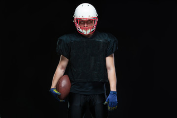 American football player in a black uniform, wearing helmet and holding ball against black background