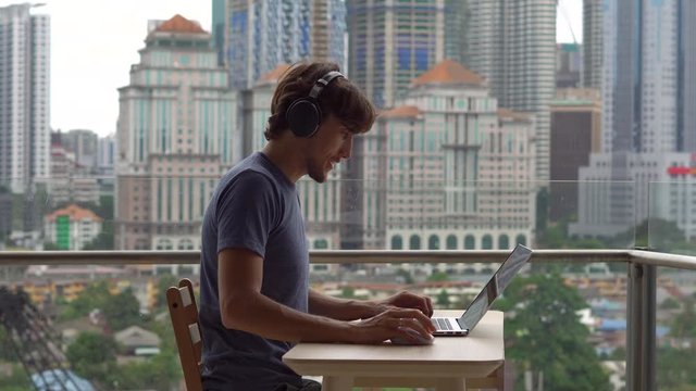 Young man freelancer workes on his laptop at a balcony with a background of a city center full of skyscrapers. Remote techer has a conversation with a student through the internet