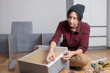 Woman assembling a chest drawer for new bedroom furniture. Female in casual shirt and knit hat does typical 