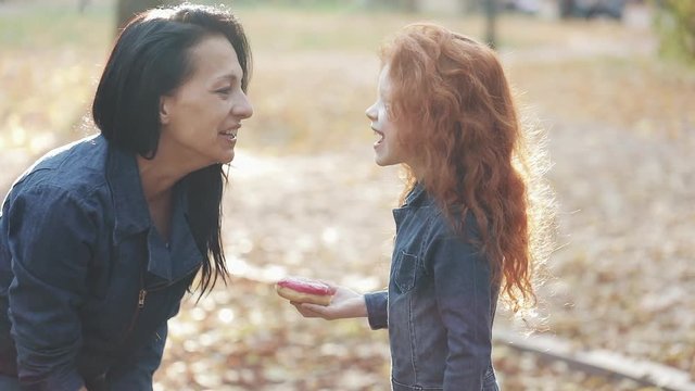 A beautiful mom and her pretty redhead daughter are standing in an autumn park. They eat donuts, feed each other, have fun