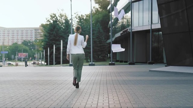 A business woman in high heels walks down the street, throws up documents, unravels her hair, removes her jacket. Freedom. Wind blows documents. Back view, slow motion.