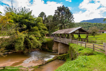 Fototapeta na wymiar Old wooden bridge covered with river, bamboo, lawn, several trees and mountains in the background, blue sky with clouds, Rio dos Cedros, Santa Catarina
