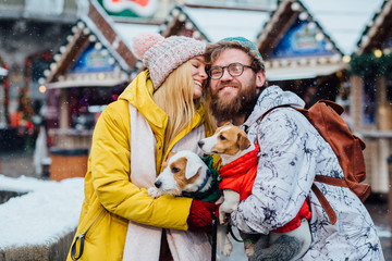 Lovely couple celebrate winter holiday conept. Happy hipster beard man and excited blond woman ,their pets Jack russel terrier hugging outdoor with Christmas market on background.