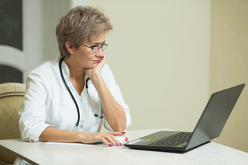 stylish aged woman in a white coat sitting at the table in the hospital with a laptop