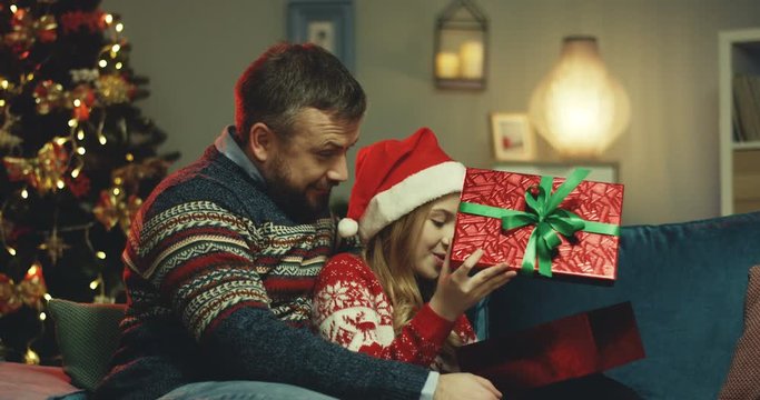 Portrait shot of the caucasian handsome father hugging his cute small daughter in the red Santa's hat while she opening a present box on the couch at the Christmas tree.