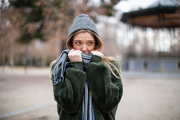 Pretty young female in warm clothes wrapping in scarf and looking away while standing on blurred background of autumn park on really cold day