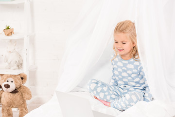 smiling child sitting on bed in pajamas and using laptop