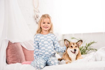 happy smiling child in pajamas sitting with corgi dog in bed