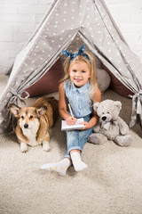 smiling child sitting with corgi dog and teddy bear in wigwam and writing in notebook