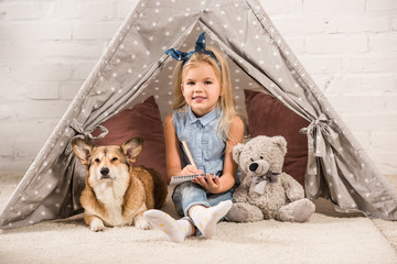 adorable child sitting with corgi dog in wigwam and writing in notebook © LIGHTFIELD STUDIOS