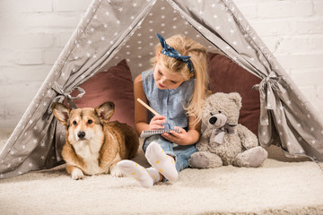 cute child sitting with corgi dog in wigwam and writing in notebook at home © LIGHTFIELD STUDIOS