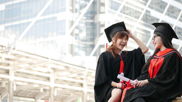 Two girls in black gowns and hold diploma certificate sitting and smiling with happy graduated.
