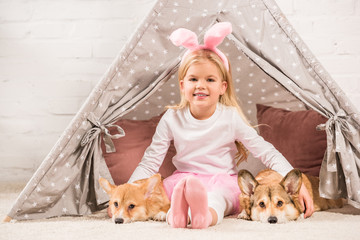 smiling child in bunny ears headband sitting with corgi dogs and teddy bear in wigwam © LIGHTFIELD STUDIOS