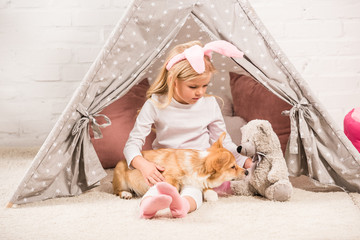 child in bunny ears headband sitting with corgi dog and teddy bear in wigwam at home