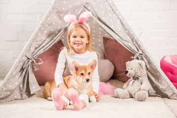 cute child in bunny ears headband sitting with corgi dog and teddy bear in wigwam © LIGHTFIELD STUDIOS