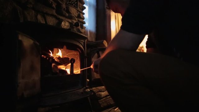 Kneeling man tends to burning fire in cast iron fireplace / wood burning stove