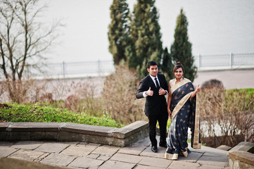 Elegant and fashionable indian friends couple of woman in saree and man in suit posed on stairs against lake.